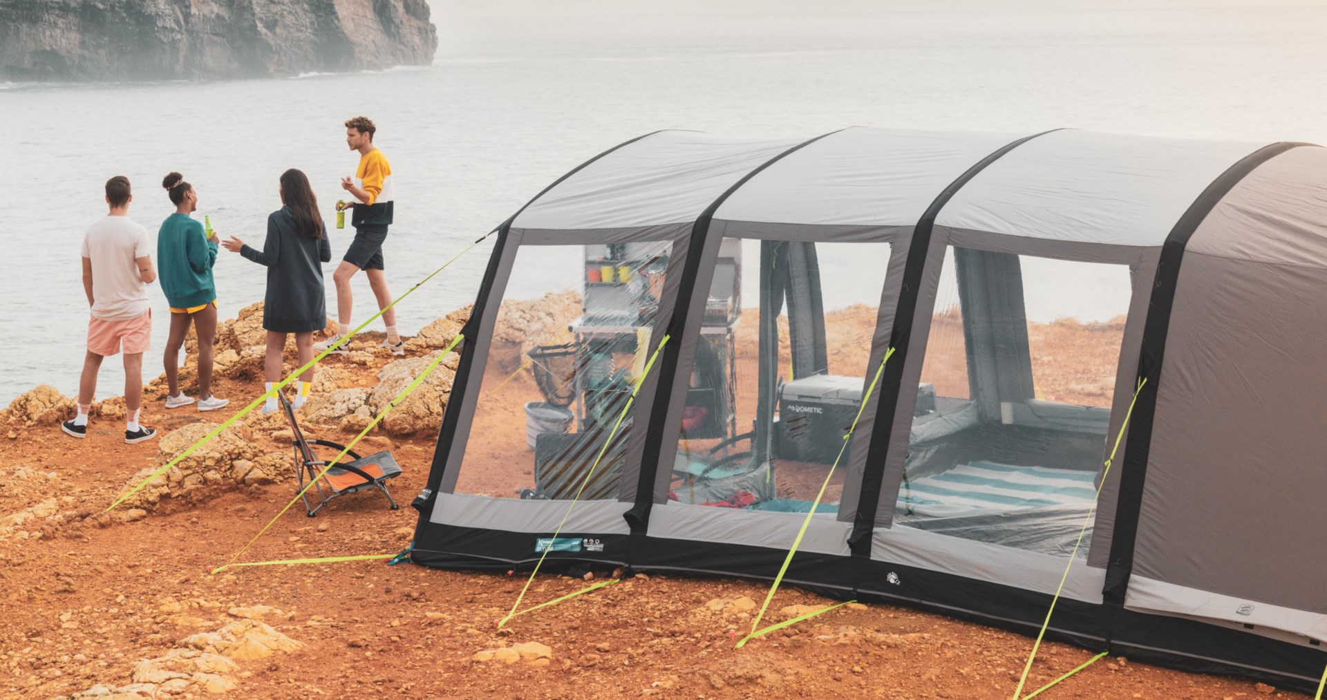 Gruppe af unge mennesker nyder socialt samvær ved campingtelt på klippekyst.